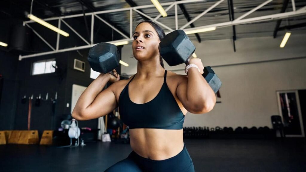 Woman in a black sports bra lifting two hex dumbbells at shoulder level during a gym workout.