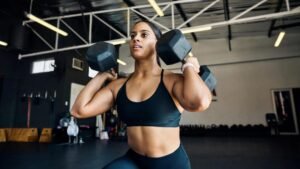 Woman in a black sports bra lifting two hex dumbbells at shoulder level during a gym workout.
