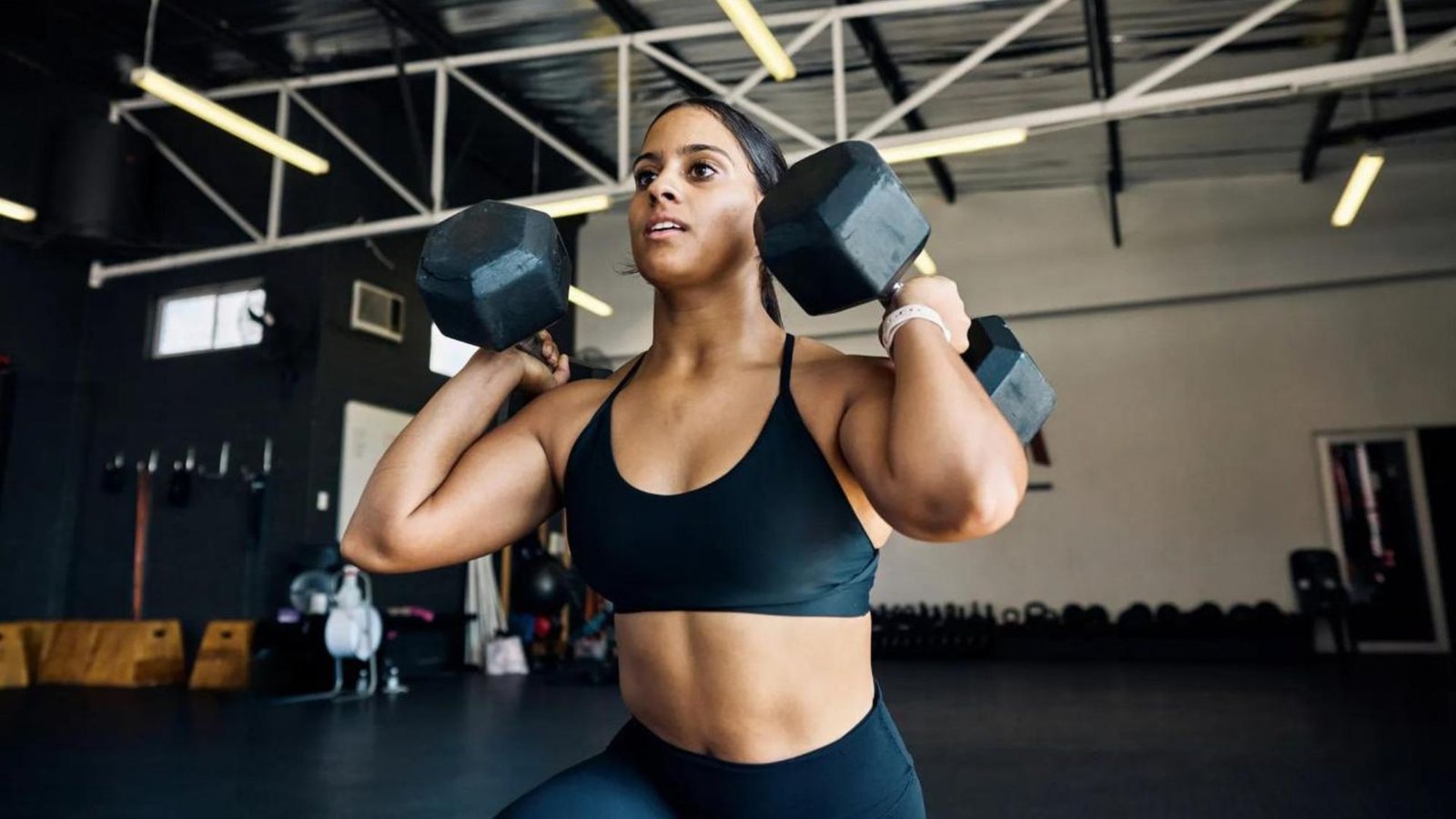 Woman in a black sports bra lifting two hex dumbbells at shoulder level during a gym workout.