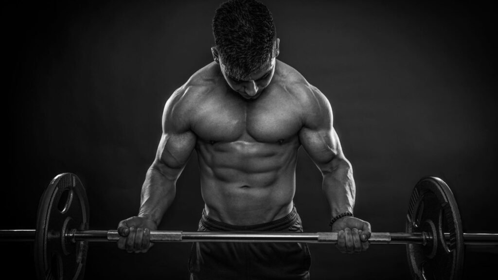 Muscular man in a dark studio lifts a loaded barbell, head down and shoulders tensed.