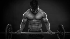 Muscular man in a dark studio lifts a loaded barbell, head down and shoulders tensed.