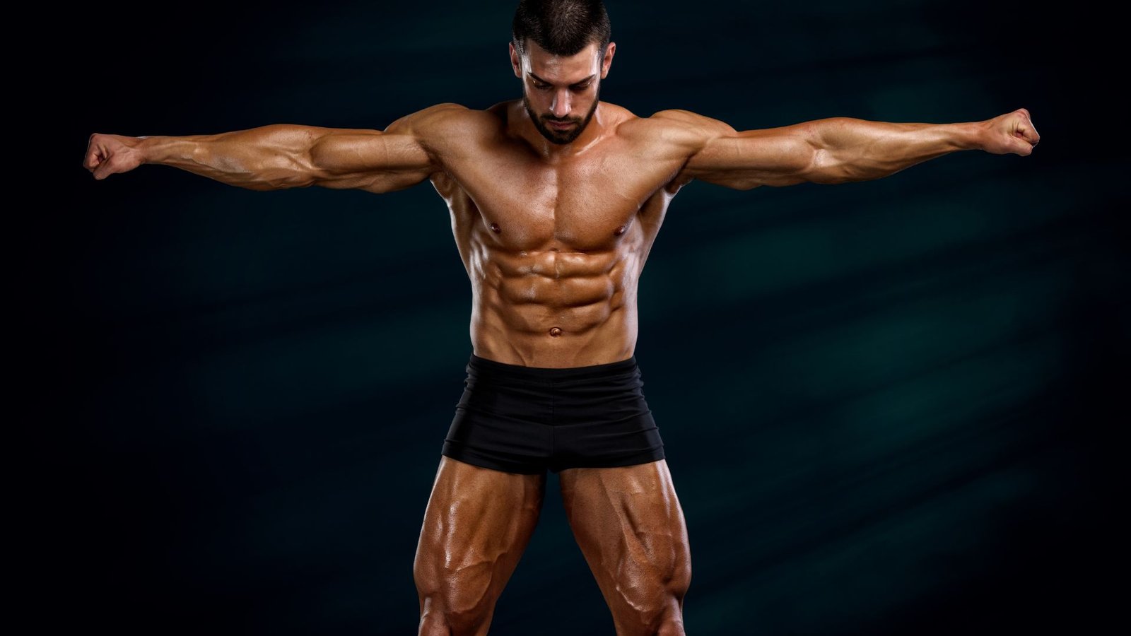 Muscular, shirtless man wearing black shorts stands with arms extended to the sides, showcasing defined abs against a dark background.
