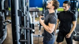 Two men in a gym: one is doing a bicep curl with a cable machine while the other watches, providing coaching.