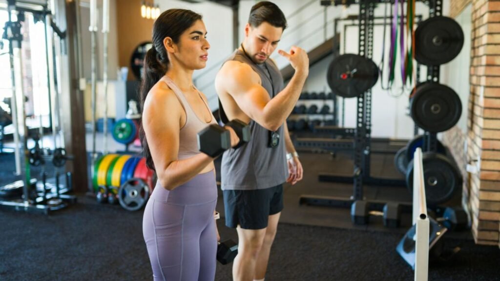 A man and a woman in a gym perform dumbbell bicep curls, with weight plates and racks in the background.