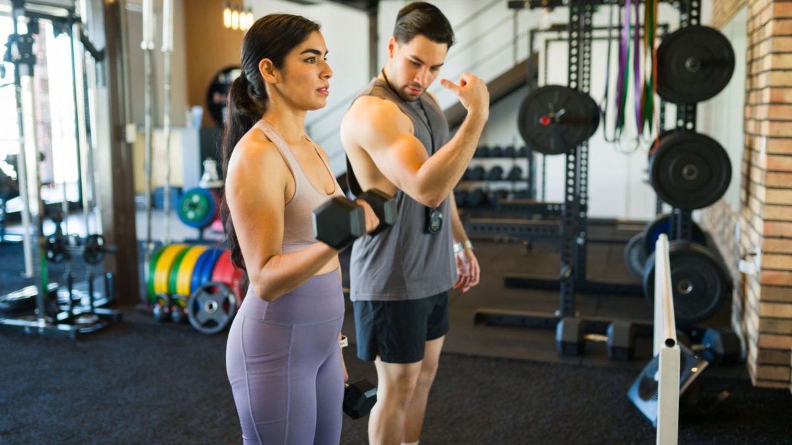 A man and a woman in a gym perform dumbbell bicep curls, with weight plates and racks in the background.