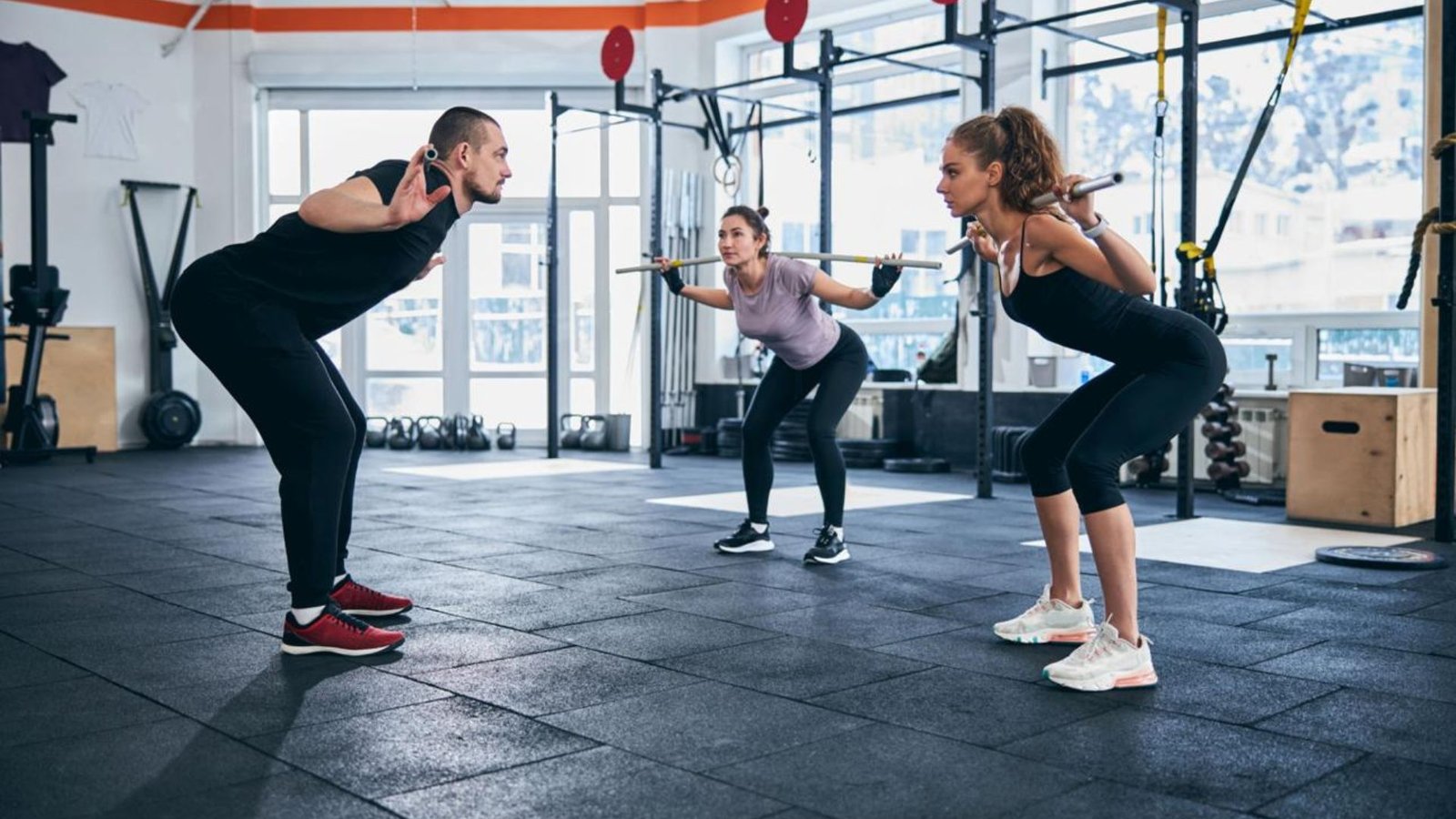 Three people in a gym performing squats with barbells, facing each other as they bend at the hips and knees.